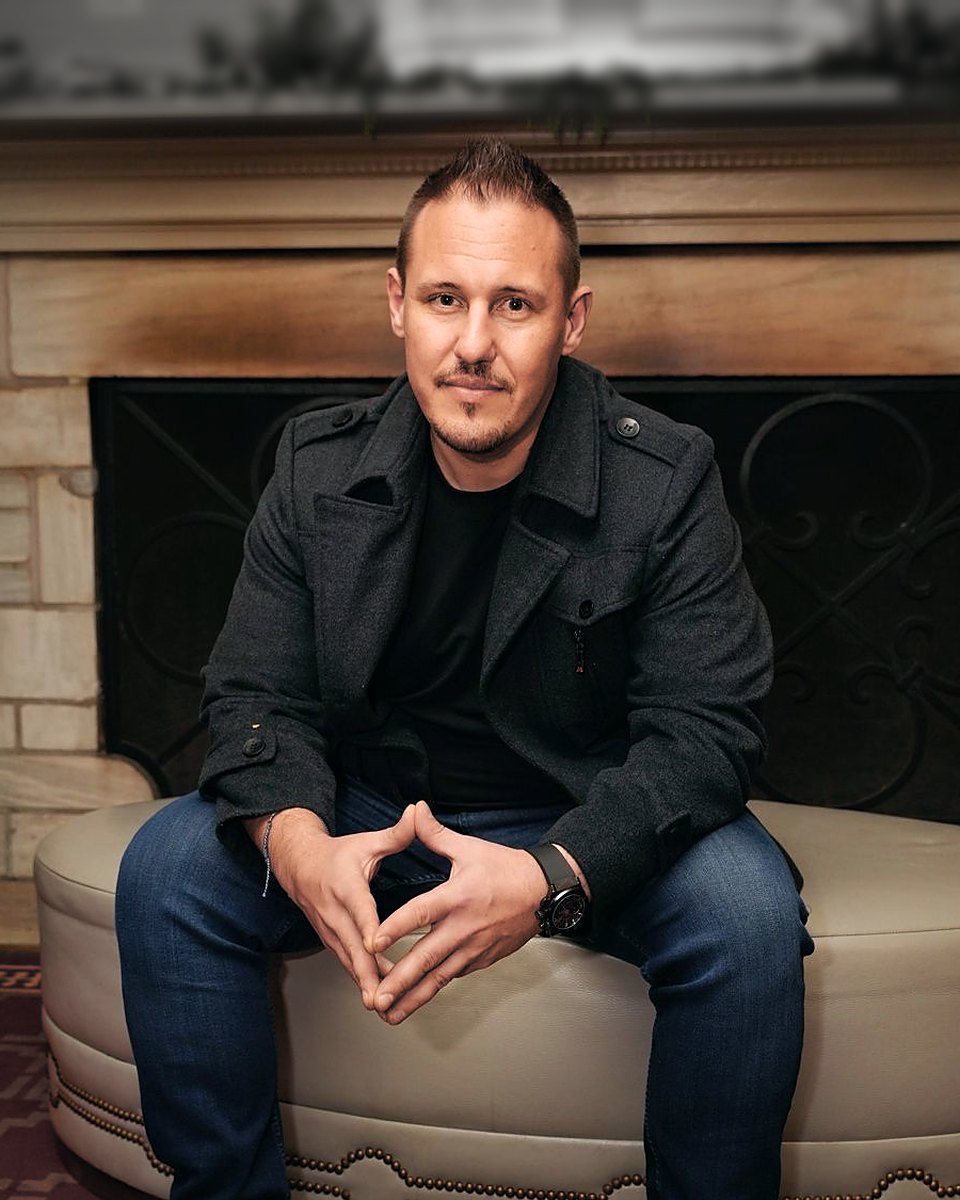 Dean DeVries, seated in front of a stone fireplace, hands clasped